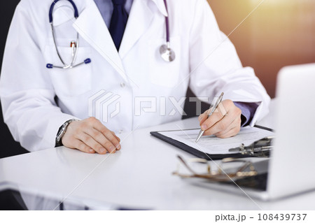 Unknown male doctor sitting and working with clipboard of medication history record in a darkened clinic, glare of light on the background, close-up of hands 108439737