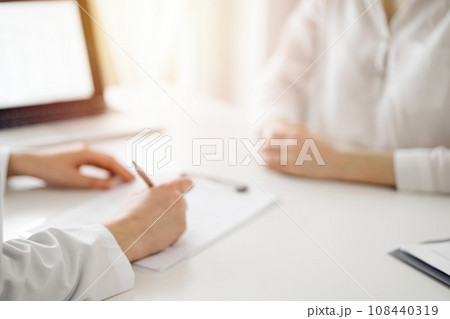 Doctor and patient sitting near each other at the white desk in clinic. Female physician is listening filling up a records form. Medicine concept 108440319