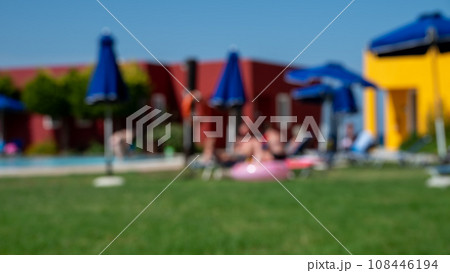Blurred shot of people relaxing and sunbathing by the pool on sun loungers and under sun umbrellas against the backdrop of bungalows. Blurred shot of people relaxing and sunbathing by the pool on sun loungers and under sun umbrellas against the backdrop of bungalows. 108446194