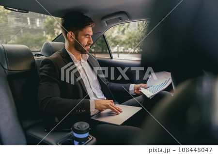 Male entrepreneur sitting on car backseat and working with documents while going to office 108448047