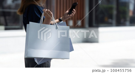 Woman using smartphone holding Black Friday shopping bag while standing on the stairs with the mall background 108455756