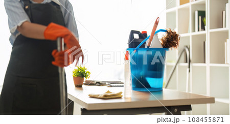 A cleaning woman is standing inside a building holding with a blue tank on the side facilities for tidying up in her hand 108455871