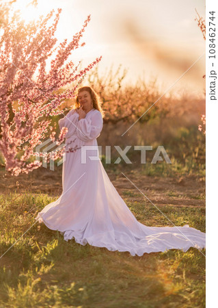 Woman blooming peach orchard. Against the backdrop of a picturesque peach orchard, a woman in a long white dress enjoys a peaceful walk in the park, surrounded by the beauty of nature. Woman blooming peach orchard. Against the backdrop of a picturesque peach orchard, a woman in a long white dress enjoys a peaceful walk in the park, surrounded by the beauty of nature. 108462744