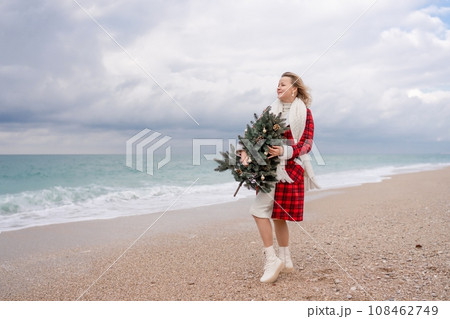Blond woman holding Christmas tree by the sea. Christmas portrait of a happy woman walking along the beach and holding a Christmas tree in her hands. Dressed in a red coat, white dress. 108462749