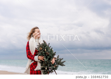 Blond woman holding Christmas tree by the sea. Christmas portrait of a happy woman walking along the beach and holding a Christmas tree in her hands. Dressed in a red coat, white dress. Blond woman holding Christmas tree by the sea. Christmas portrait of a happy woman walking along the beach and holding a Christmas tree in her hands. Dressed in a red coat, white dress. 108462750