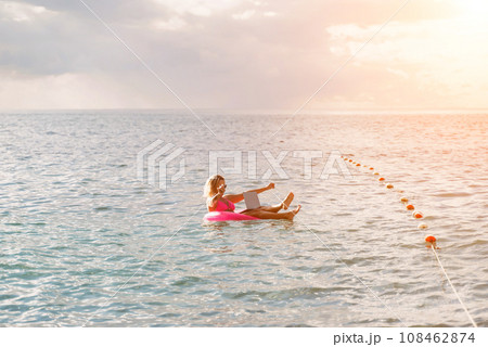 Woman works on laptop in sea. Freelancer, blond woman in sunglases floating on an inflatable big pink donut with a laptop in the sea at sunset. People summer vacation rest lifestyle concept. Woman works on laptop in sea. Freelancer, blond woman in sunglases floating on an inflatable big pink donut with a laptop in the sea at sunset. People summer vacation rest lifestyle concept. 108462874