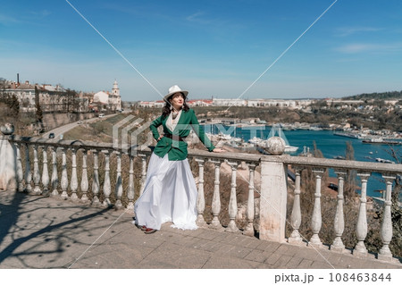Woman walks around the city, lifestyle. Happy woman in a green jacket, white skirt and hat is sitting on a white fence with balusters overlooking the sea bay and the city. 108463844