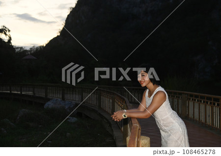 Portrait photography of a mature, beautiful, happy Asian woman. Model in white casual clothes. Standing and smiling at the camera, standing on a bridge with mountains behind. Portrait photography of a mature, beautiful, happy Asian woman. Model in white casual clothes. Standing and smiling at the camera, standing on a bridge with mountains behind. 108465278