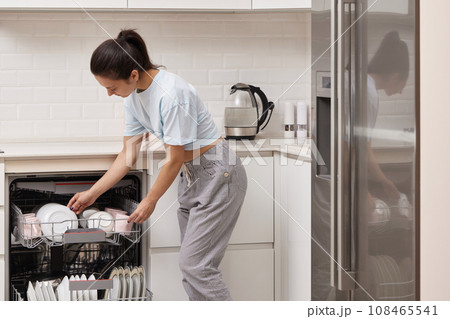 happy woman unloading plate from open automatic built-in dishwasher machine with clean utensils inside in modern white kitchen. happy woman unloading plate from open automatic built-in dishwasher machine with clean utensils inside in modern white kitchen. 108465541
