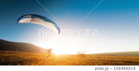 A paraglider glides along the ground at sunset with mountains in the background. Panoramic shot banner for paragliding in warm colors. Glare from the sun in the frame A paraglider glides along the ground at sunset with mountains in the background. Panoramic shot banner for paragliding in warm colors. Glare from the sun in the frame 108466018