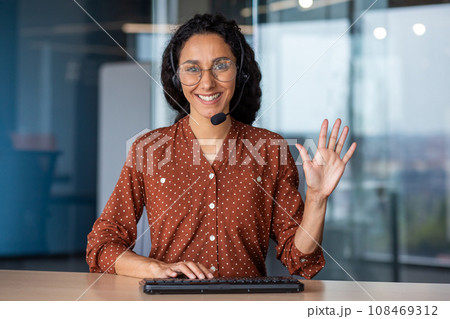 Portrait of young successful Latin American online support worker, woman smiling and looking at camera with headset phone, webcam view, typing on keyboard, inside office at workplace. 108469312
