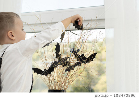 Halloween concept. Autumn holiday. The boy stands with his back near the window against the backdrop of a black vase with batting mice cut out of paper on dry branches. Close-up. 108470896