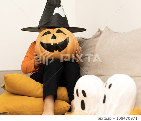 Halloween. Concept. A boy in an orange sweater and a haunted wizard's hat, with a pumpkin instead of a face, sits cheerfully in a home interior. Halloween. Concept. A boy in an orange sweater and a haunted wizard's hat, with a pumpkin instead of a face, sits cheerfully in a home interior. 108470971