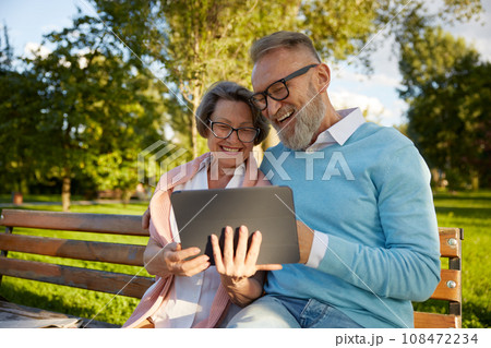 Happy cheerful senior couple using digital tablet while rest on park bench Happy cheerful senior couple using digital tablet while rest on park bench 108472234
