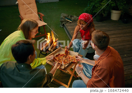 Happy friends enjoying pizza and beer drink nearby burning campfire 108472440
