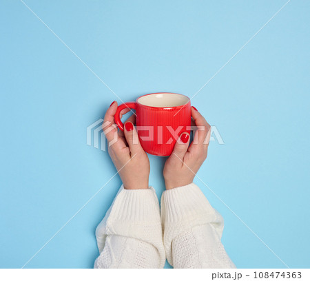 Two female hands holding a red ceramic cup on a blue background Two female hands holding a red ceramic cup on a blue background 108474363