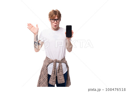 young surprised european red-haired guy in a t-shirt shows a vertical smartphone 108476518