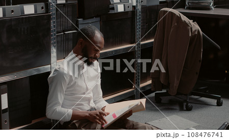 Police officer sitting on archive room floor with files conducting investigation, having piles of evidence and records in folders. Detective gathering case information in incident room. Police officer sitting on archive room floor with files conducting investigation, having piles of evidence and records in folders. Detective gathering case information in incident room. 108476741