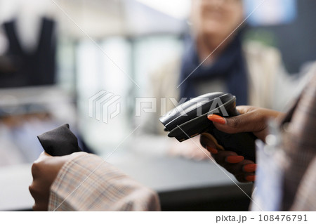 African american worker scanning fashionable shirt using store scanner, preparing purchase for elderly shopaholic woman. Client buying casual wear in clothing store, commercial activity. Close up African american worker scanning fashionable shirt using store scanner, preparing purchase for elderly shopaholic woman. Client buying casual wear in clothing store, commercial activity. Close up 108476791