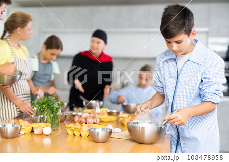During cooking classes, teen boy carefully mixes ingredients in bowl with whisk 108478958