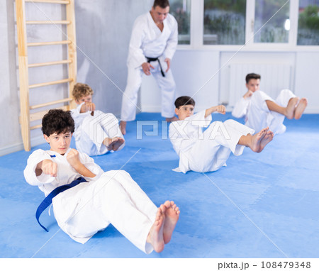 Concentrated boys in kimono doing abdominal exercise and practicing punches in gym during group martial arts workout 108479348