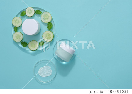 Against the light blue background, cosmetic jars without label decorated with fresh slices of cucumber and mint leaves on transparent podiums. Mockup for design, advertising photo for cosmetic 108491031