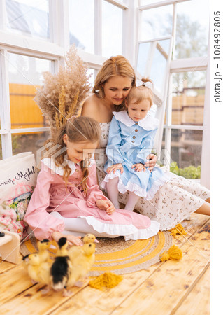 Vertical shot of cheerful young mom and two adorable daughters in dress playing with little yellow ducklings in summer gazebo house on sunny day. Concept of excursion to eco-farm, life in village. Vertical shot of cheerful young mom and two adorable daughters in dress playing with little yellow ducklings in summer gazebo house on sunny day. Concept of excursion to eco-farm, life in village. 108492806