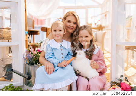 Portrait of cute mother and two adorable daughters in dress hugging white rabbit in summer gazebo house on sunny day, smiling looking at camera. Concept of excursion to eco-farm, life in village. 108492814