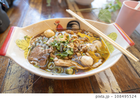 Thai noodle with pork, meatball, pork liver with blood soup in bowl that look like boat on the wood table. Thai noodle with pork, meatball, pork liver with blood soup in bowl that look like boat on the wood table. 108495524