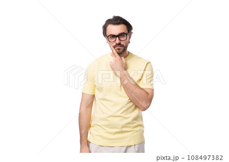 a young European brunette guy with a beard and mustache in glasses stands thoughtfully on a white 108497382