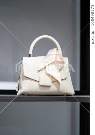 White cream handbag for women with cream ribbon tied on the handle of the bag is placed on a wooden shelf. White cream handbag for women with cream ribbon tied on the handle of the bag is placed on a wooden shelf. 108498375