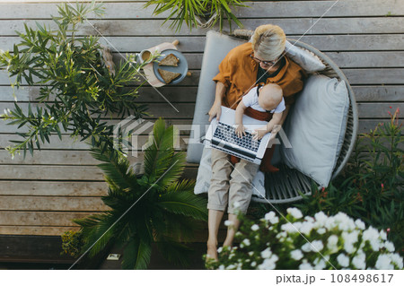Top view of mother holding baby while working on laptop in garden. Businesswoman working remotely from outdoor home office and taking care of little son. Life work balance with kid. Top view of mother holding baby while working on laptop in garden. Businesswoman working remotely from outdoor home office and taking care of little son. Life work balance with kid. 108498617