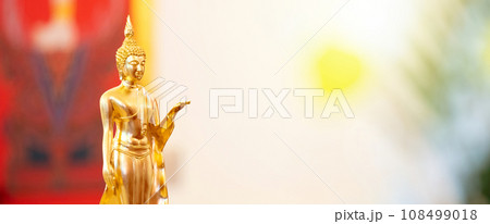 Standing Buddha statue is placed on top of a rectangular object in front of a red framed picture and tree house. Standing Buddha statue is placed on top of a rectangular object in front of a red framed picture and tree house. 108499018