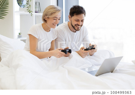 Caucasian playing video games while lying under blanket at home. 108499363