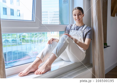 Portrait of smiling teenage girl sitting on windowsill at home 108500195