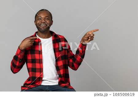 Positive black bearded man wearing red checkered shirt pointing at advertisement area Positive black bearded man wearing red checkered shirt pointing at advertisement area 108500619