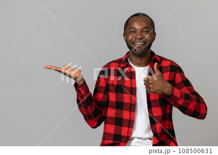 Joyful black bearded man wearing red checkered shirt presenting copy space on palm showing thumb up 108500631