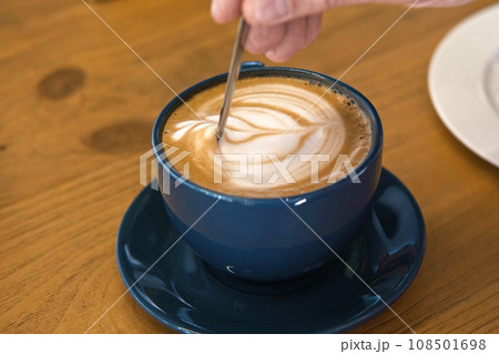 Hand is Mixing the Milk Foam of Latte. Person Stirring Cappuccino with Steel Spoon. Hot Coffee in a White Classic Ceramic Cup on Wooden Table. Modern Coffee shop, Cafeteria. Thick Froth. Close-up View 108501698