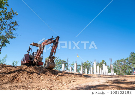Hydraulic excavator is digging soil at a construction site, surrounded by lot of trees, under clear sky. Clawer backhoe parked under tree shade. 108502198