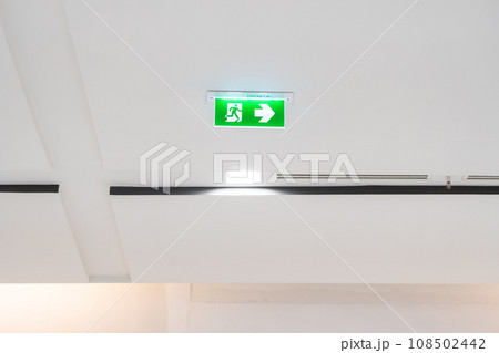 White neon sign depicting a fire exit with green background hangs from the ceiling. White neon sign depicting a fire exit with green background hangs from the ceiling. 108502442