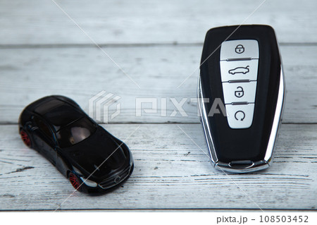 The car keys are black with metal inserts and automatic buttons lying on a background of white wood, next to them is a black car close-up. High quality photo The car keys are black with metal inserts and automatic buttons lying on a background of white wood, next to them is a black car close-up. High quality photo 108503452
