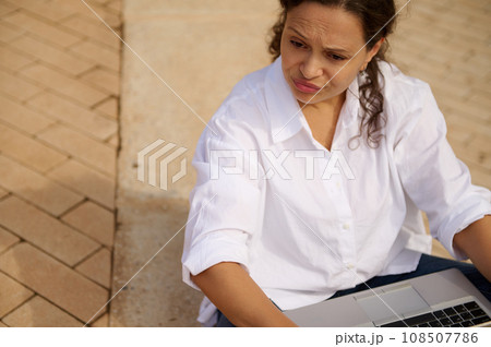 Mixed race young woman expressing sadness failing business tender project, looking aside, sitting with laptop outdoors Mixed race young woman expressing sadness failing business tender project, looking aside, sitting with laptop outdoors 108507786