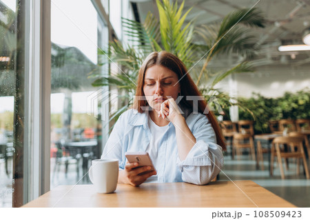 Portrait of sad young redhead woman in a coffee shop looking at smart phone. A lonely unhappy 30s girl siting in a cafe. Bad feelings stress, anxiety, grief, emotions. Portrait of sad young redhead woman in a coffee shop looking at smart phone. A lonely unhappy 30s girl siting in a cafe. Bad feelings stress, anxiety, grief, emotions. 108509423