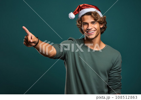 Young man in Santa hat, smiling guy pointing finger to side. Portrait of handsome person isolated on blue green studio background. Concept of Christmas, show, welcome, xmas, gif 108512863