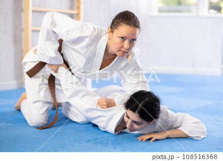 Sportive woman holds her opponent by force while sitting on mats during judo classes 108516368