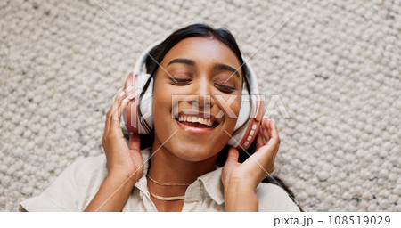 Headphones, happy and young woman on the carpet on the floor listening to music or album. Smile, calm and Indian female person streaming podcast, radio or playlist and chilling in living room of home Headphones, happy and young woman on the carpet on the floor listening to music or album. Smile, calm and Indian female person streaming podcast, radio or playlist and chilling in living room of home 108519029