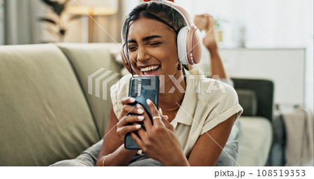Woman, singing and phone with headphones on sofa, smile or relax for comic performance in living room. Girl, smartphone and audio streaming subscription with voice, noise or karaoke on couch in house 108519353