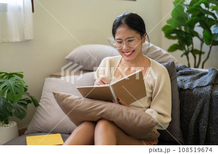 A happy Asian woman is sitting on the floor beside her bed in the bedroom and keeping her diary. 108519674