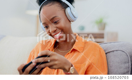 Smartphone games, headphones or woman in home on sofa for playing online subscription, digital gaming or connection. Happy african person, video game or mobile in living room for streaming multimedia 108520703