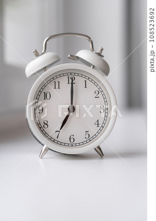 Close-up of a round white alarm clock on the windowsill in the bedroom. The hands on the clock show seven o'clock in the morning, time to get up. Retro alarm clock on the table. space for text Close-up of a round white alarm clock on the windowsill in the bedroom. The hands on the clock show seven o'clock in the morning, time to get up. Retro alarm clock on the table. space for text 108523692